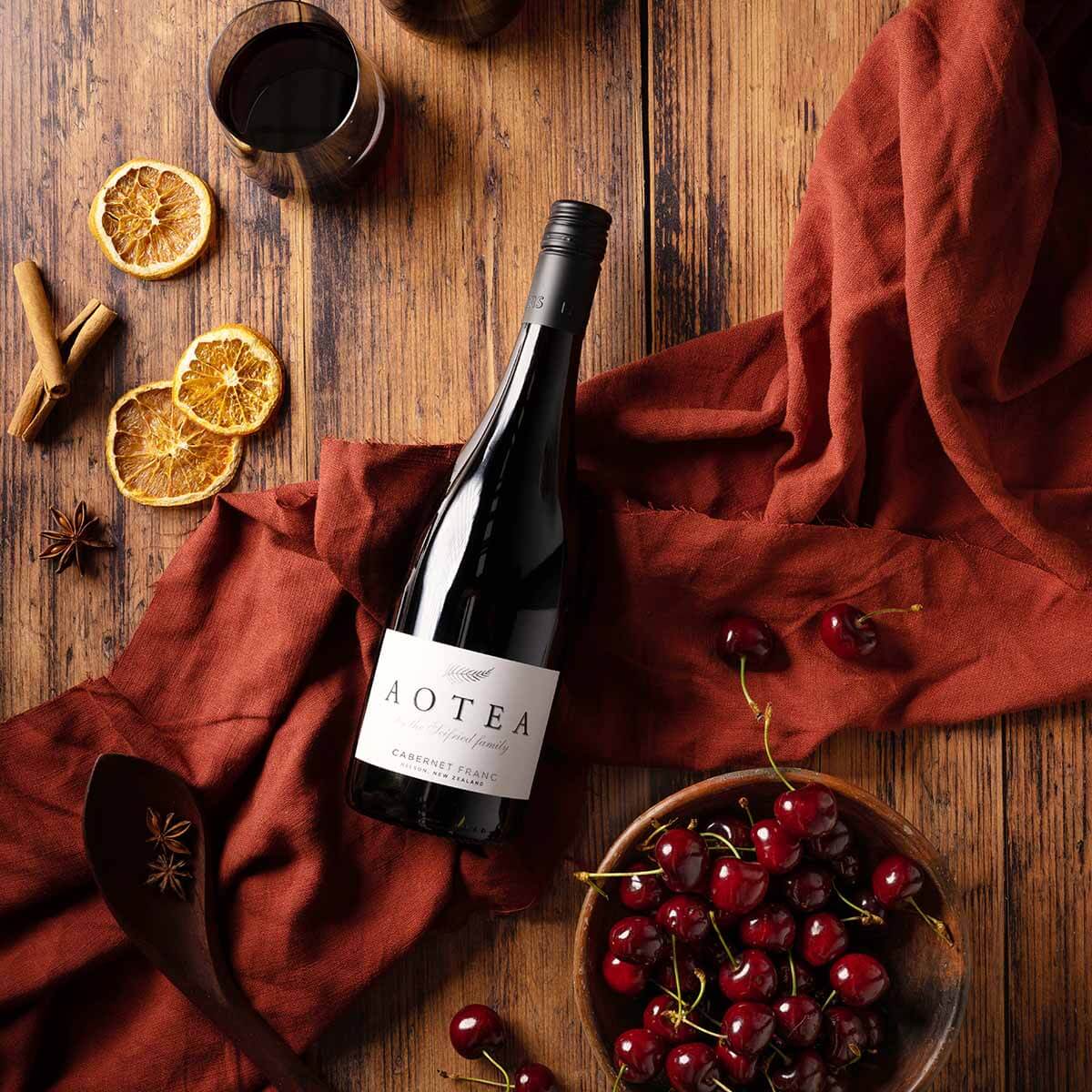 Flatlay bottle of Aotea by the Seifried family Nelson Cabernet Franc on a rust-orange cloth with a glass of red wine, fresh cherries, and spices