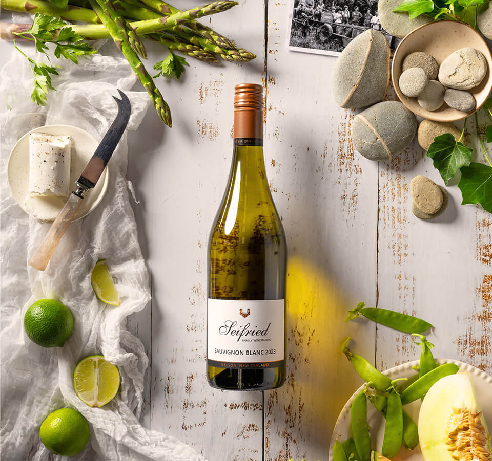 Top-down view of Old Label Seifried Nelson Sauvignon Blanc bottle styled with limes, goat cheese, fresh peas, melon, and river stones