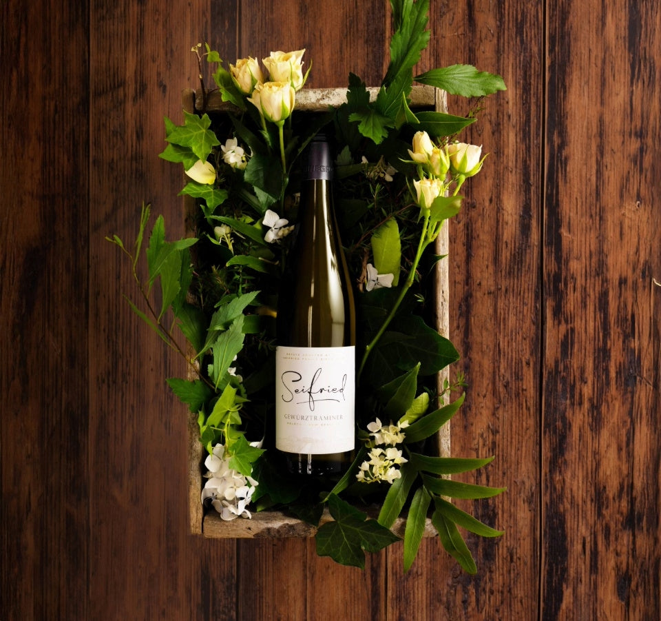 Bottle of Seifried Gewürztraminer nestled in green foliage and white flowers inside a wooden crate against a dark wood backdrop