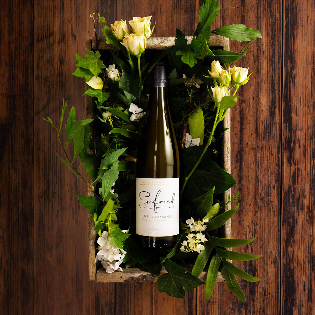 Bottle of Seifried Gewürztraminer wine nestled in green foliage and white flowers inside a wooden box against a dark wood backdrop