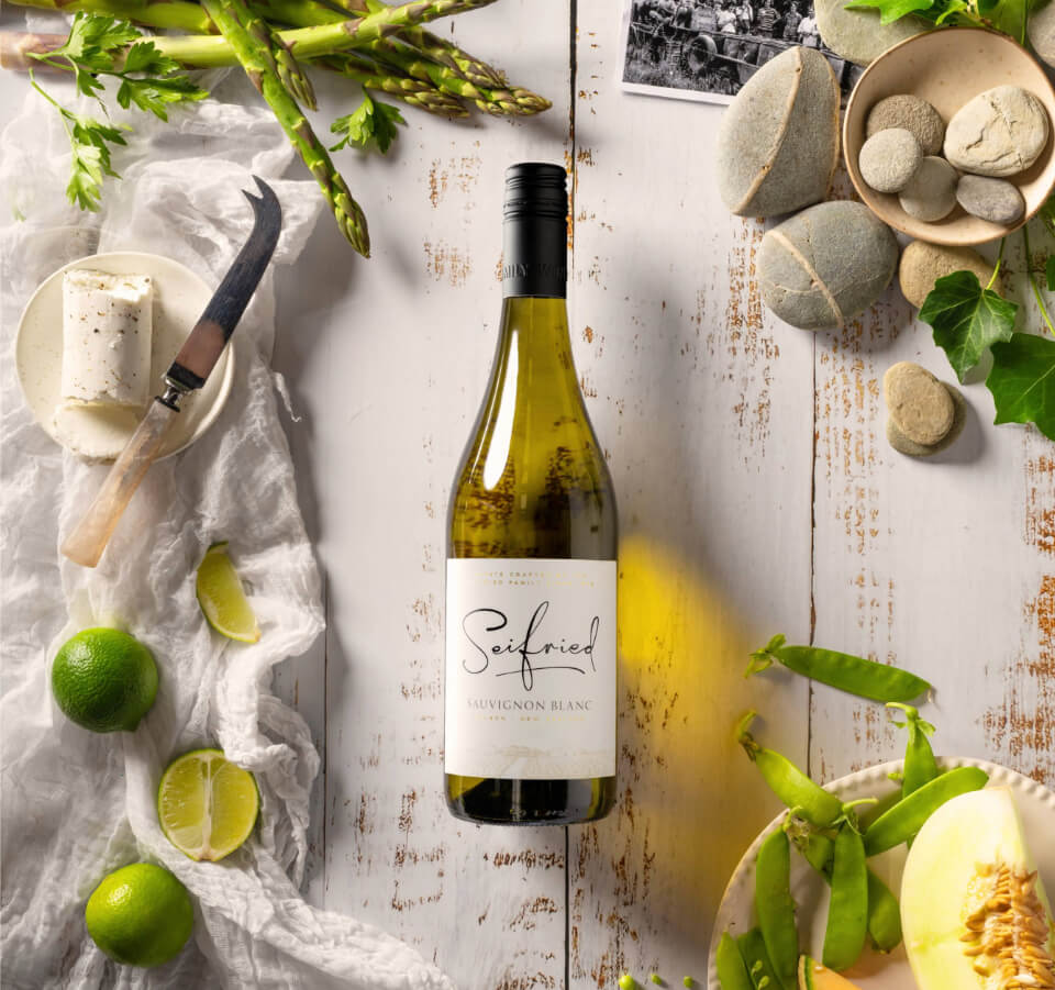 Bottle of Seifried Sauvignon Blanc on a white wooden table surrounded by limes, cheese, river stones, and fresh produce